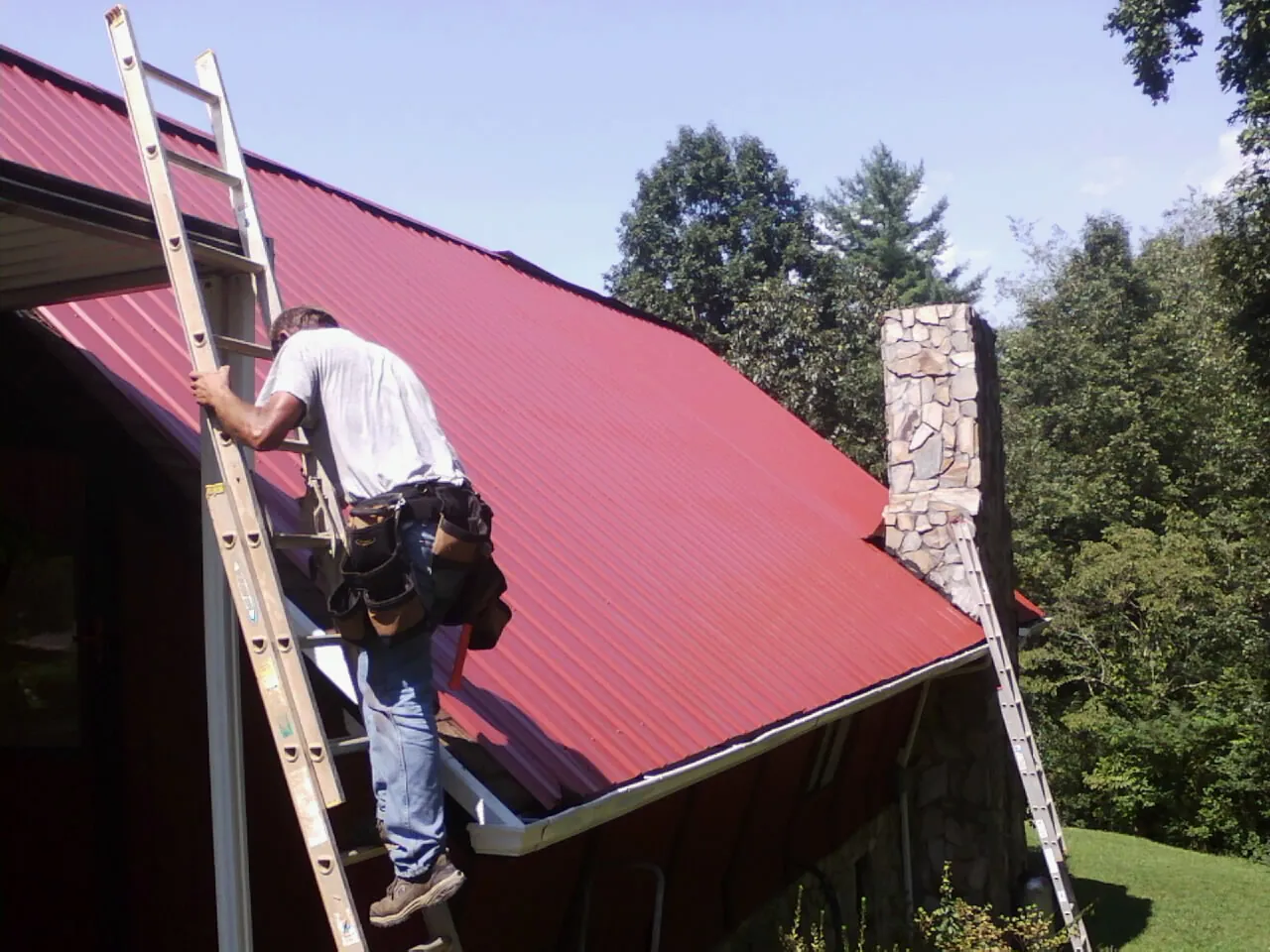 Roofer on a ladder beside a red metal roof and stone chimney — Soffit & Fascia Repair by Roofing Brothers in Vanderbilt